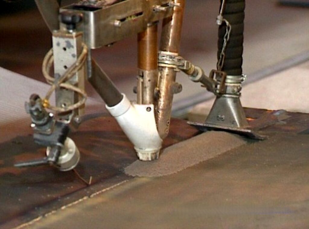 Tête de soudage SAW mécanisée sur une virole en acier, avec flux granulaire couvrant le joint.